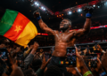 Francis Ngannou célèbre une victoire spectaculaire dans une arène de MMA bondée, entouré de supporters filmant la scène avec leurs smartphones tandis qu’un drapeau camerounais flotte au premier plan.