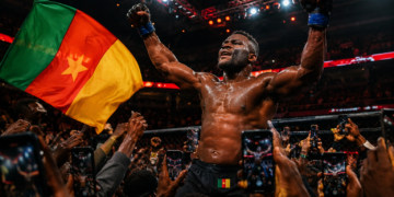 Francis Ngannou célèbre une victoire spectaculaire dans une arène de MMA bondée, entouré de supporters filmant la scène avec leurs smartphones tandis qu’un drapeau camerounais flotte au premier plan.