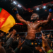 Francis Ngannou célèbre une victoire spectaculaire dans une arène de MMA bondée, entouré de supporters filmant la scène avec leurs smartphones tandis qu’un drapeau camerounais flotte au premier plan.