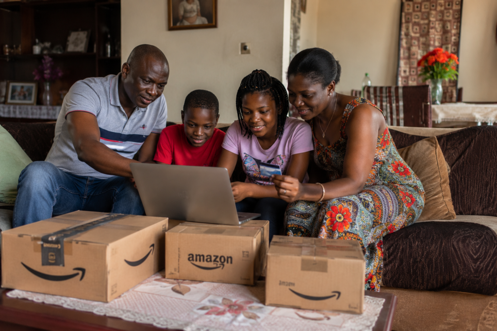 Famille camerounaise réunie dans son salon autour d'un ordinateur portable et de smartphones pour effectuer des achats sur Amazon, avec plusieurs colis de livraison visibles au premier plan.
