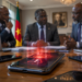 Illustration d'un smartphone affichant une alerte de surveillance au premier plan, tandis que des responsables télécoms et gouvernementaux discutent lors d'une réunion sur le blocage des téléphones non dédouanés au Cameroun.