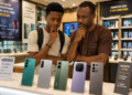 Un parent et son enfant dans une boutique de téléphonie mobile examinent cinq smartphones Android exposés sur un comptoir, parmi lesquels le TECNO Camon 50 Pro, le Samsung Galaxy A56 5G, le POCO X7 Pro, le Redmi Note 14 Pro+ 5G et l'Infinix Note 50 Pro.