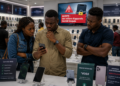 Contrôle IMEI : Orange et MTN sommés de suspendre les appareils non dédouanés 70 Des clients africains consultent un smartphone dans une boutique de téléphonie moderne tandis qu'un écran d'alerte lié au contrôle des IMEI est visible en arrière-plan.