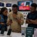 Des clients africains consultent un smartphone dans une boutique de téléphonie moderne tandis qu'un écran d'alerte lié au contrôle des IMEI est visible en arrière-plan.