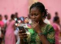 Une jeune femme camerounaise pensive regarde son smartphone, entourée d'icônes de réseaux sociaux en lévitation affichant un démenti sur la mort du père de Lionel Messi avec le portrait du joueur.