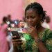 Une jeune femme camerounaise pensive regarde son smartphone, entourée d'icônes de réseaux sociaux en lévitation affichant un démenti sur la mort du père de Lionel Messi avec le portrait du joueur.