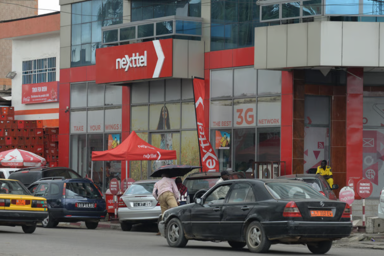 Façade d’un bâtiment aux couleurs de Nexttel Cameroun, avec l’enseigne de l’opérateur mobile, des véhicules stationnés et des installations commerciales.